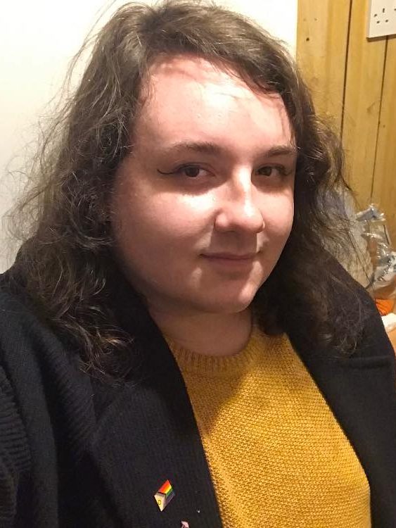 A young white transgender woman with curly, shoulder-length brown hair smiles into the camera. She's wearing a small amount of eye makeup, a long blue coat and a mustard yellow jumper underneath. On the left lapel of the coat is a metal pin styled after the Progress Pride Flag. She appears to be in a kitchen. The pain in her eyes suggests she is British.
